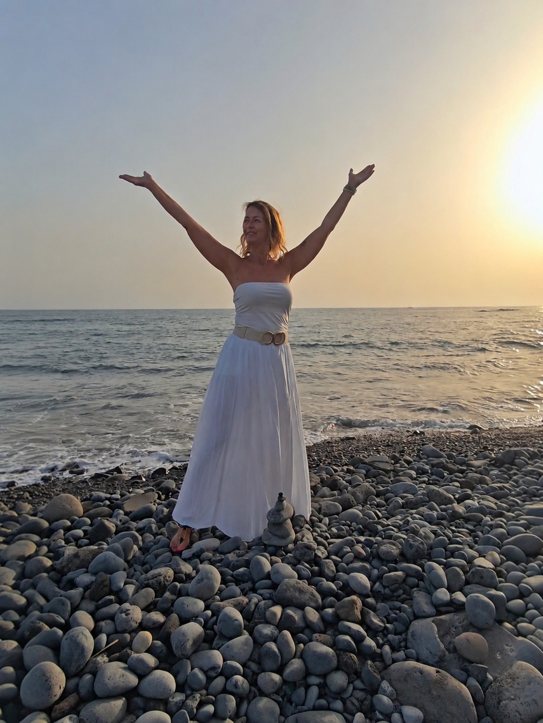 Mathilda in witte kleding aan zee op Tenerife, met haar armen omhoog in het avondlicht.